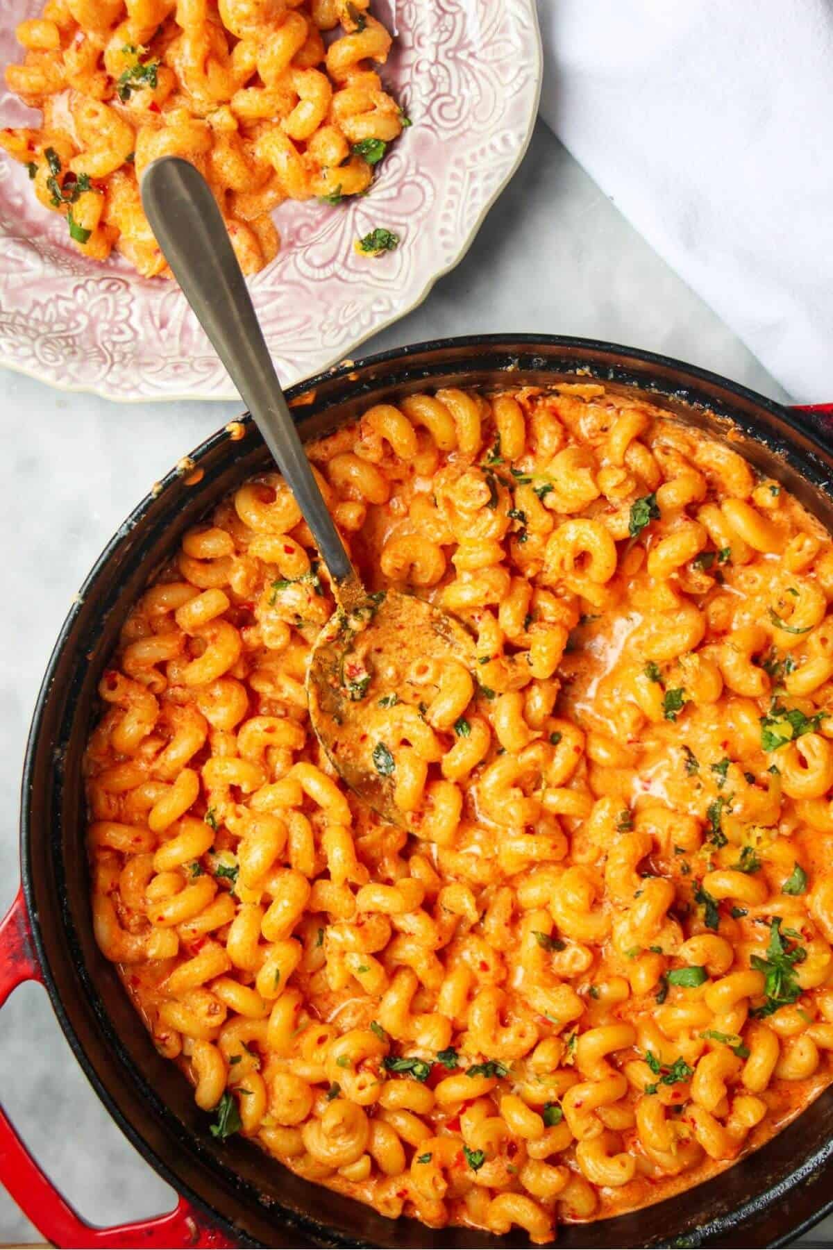 Spicy mac and cheese in a large black skillet with a silver spoon inside and pink plate next to it.