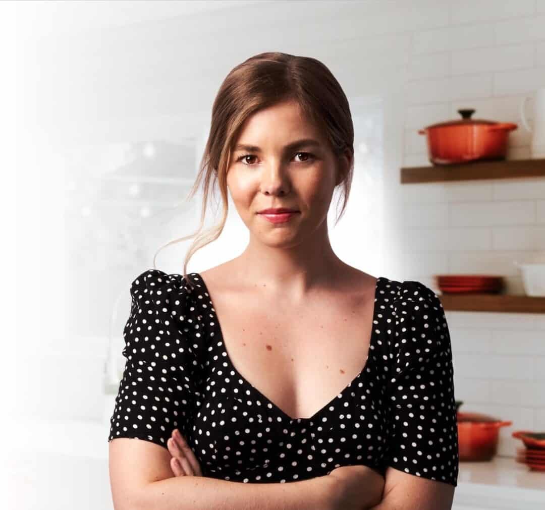Kate in a kitchen with red pots in the background with arms folded.