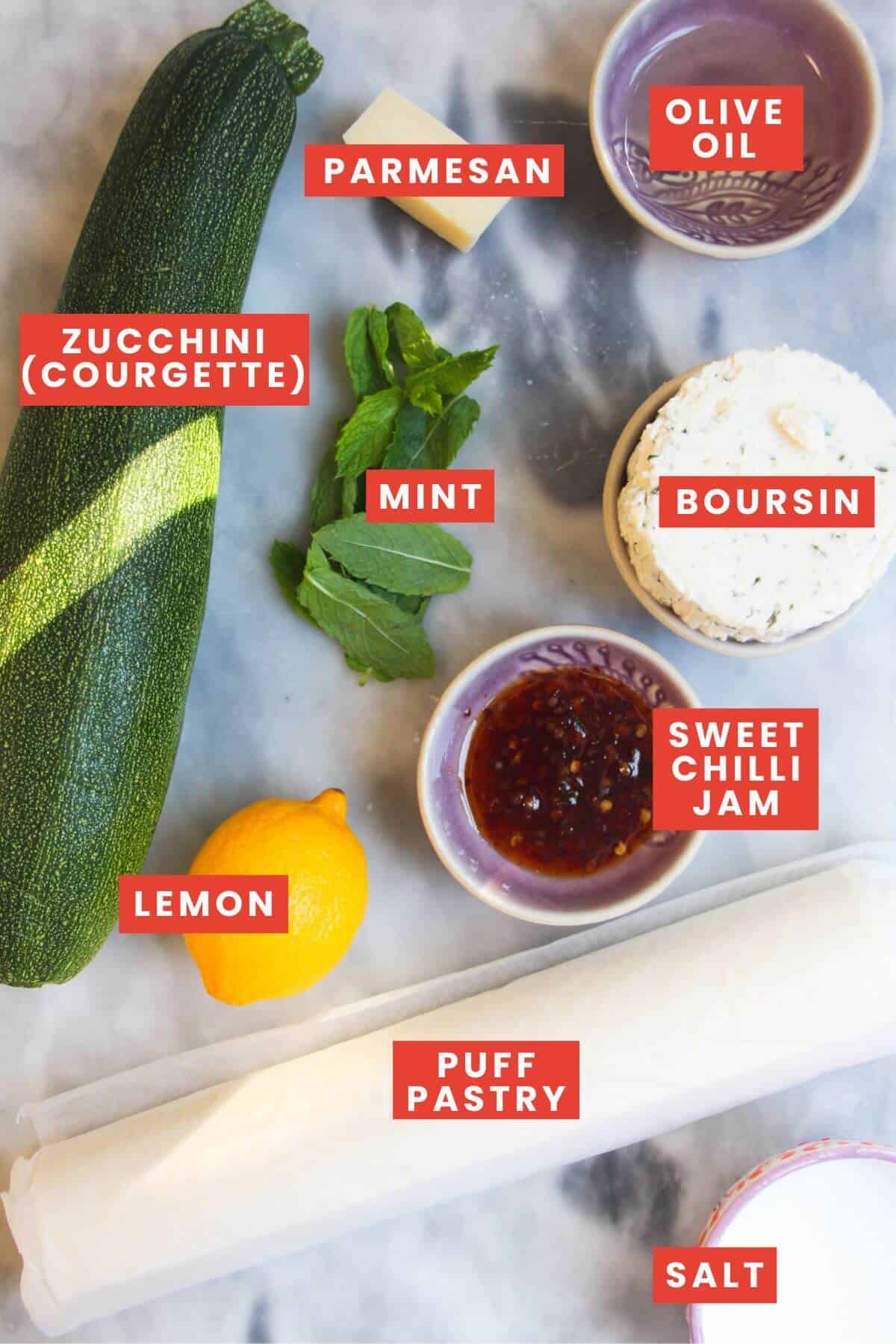 Ingredients for zucchini puff pastry tart laid out on a grey marble background and labelled.
