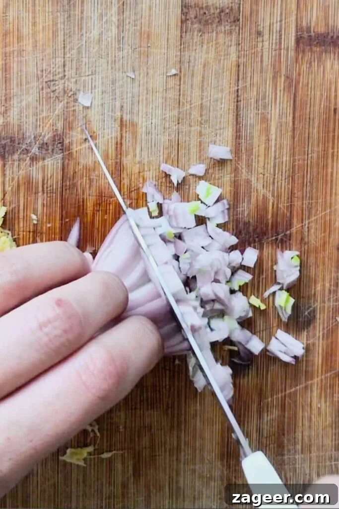 Hand with large knife chopping shallot on a wooden board.