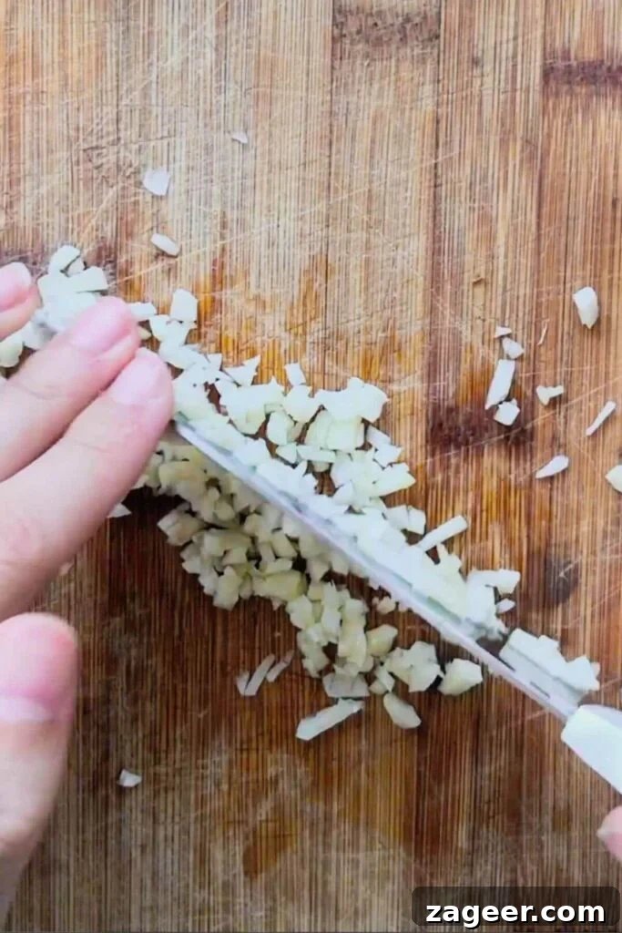 Hand with knife chopping garlic finely on a wooden board.