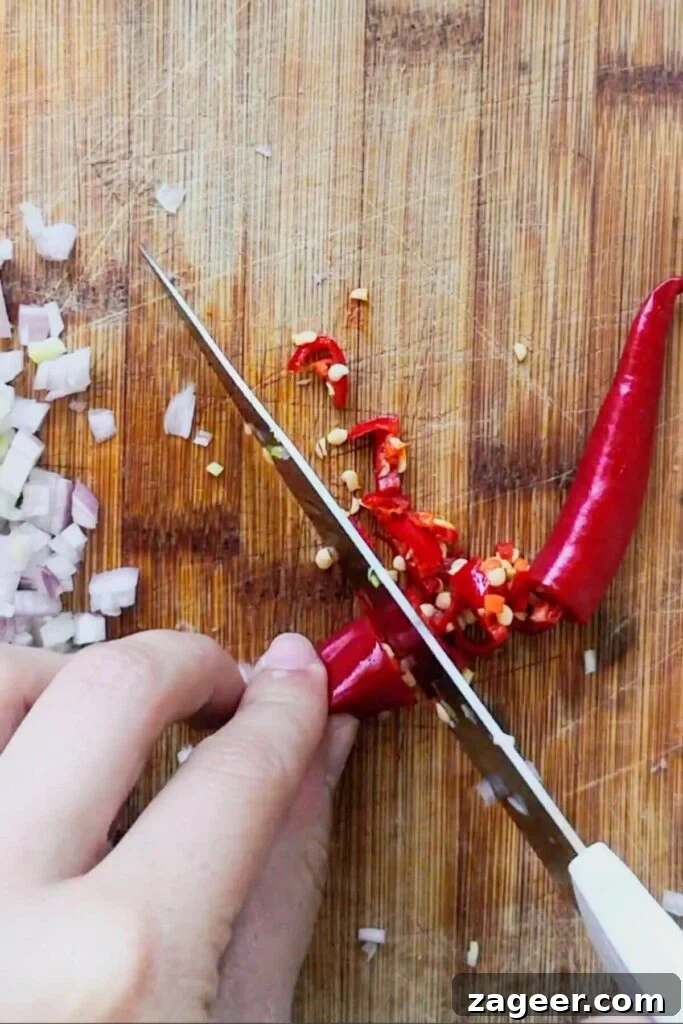 Hand with knife chopping red chili on a wooden board.