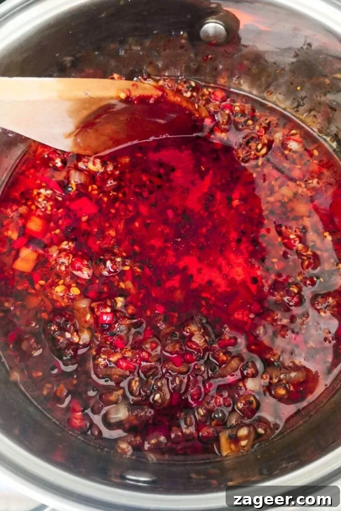Chili oil simmering gently in a small silver pot with a wooden spoon inside.