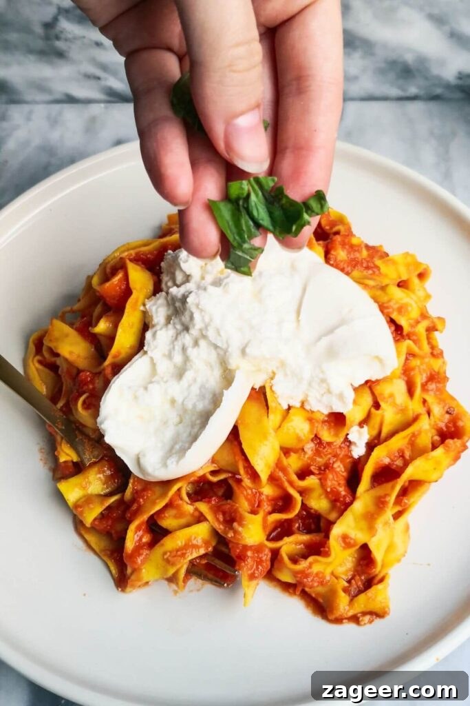 Quick Spicy Nduja Pasta with Creamy Burrata 9 A hand adding fresh basil to burrata on top of nduja pasta on a large white plate for garnish.