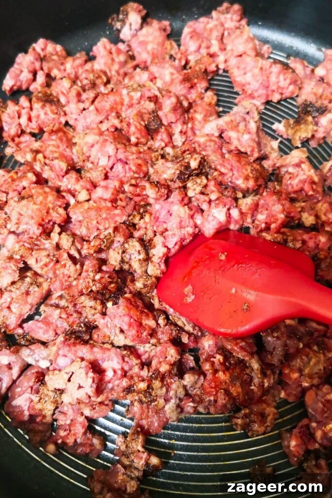 Sticky Beef Fatteh Middle Eastern Layered Nachos 4 Ground beef being stirred with a large red spatula in a large pan, mixing with fragrant spices.