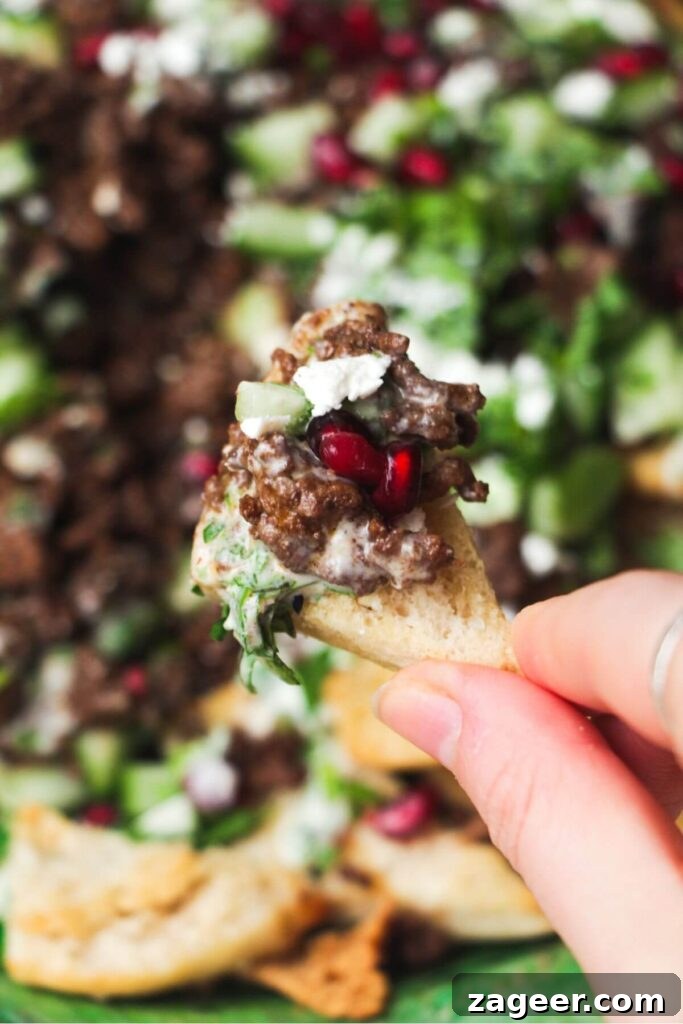 Sticky Beef Fatteh Middle Eastern Layered Nachos 7 A close-up shot of a hand holding a loaded pita chip, topped with sticky beef, vibrant pomegranate seeds, and fresh herbs, highlighting the delicious textures.
