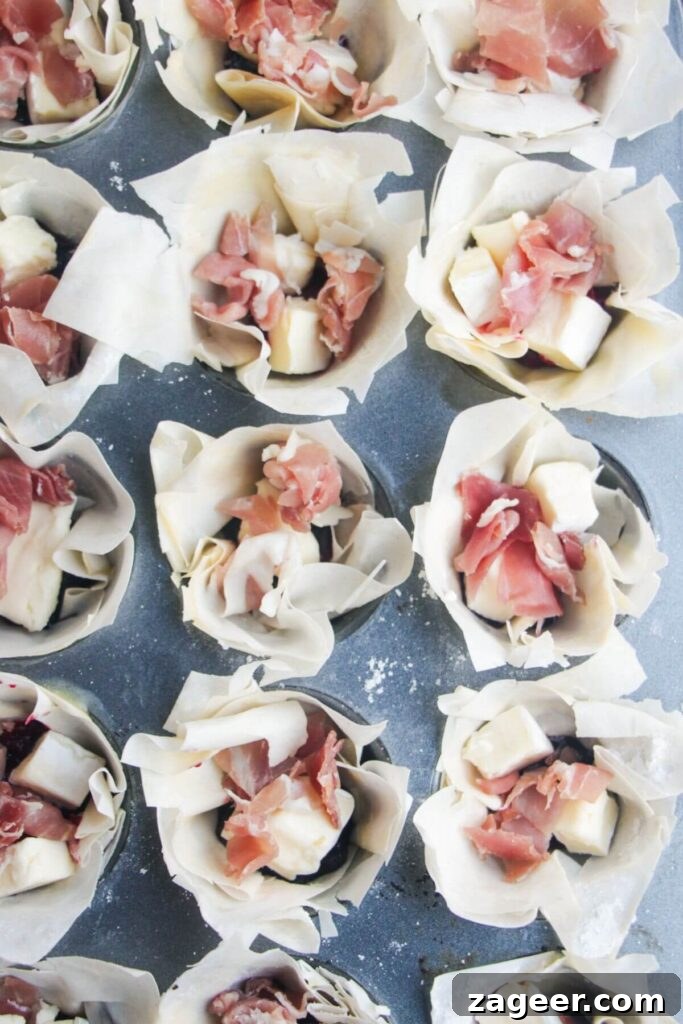 Effortless Brie, Cranberry, and Prosciutto Filo Parcels 6 Mini filo cups generously filled with a dollop of vibrant cranberry sauce, small pieces of savory prosciutto, and creamy cubes of brie cheese, ready for baking.