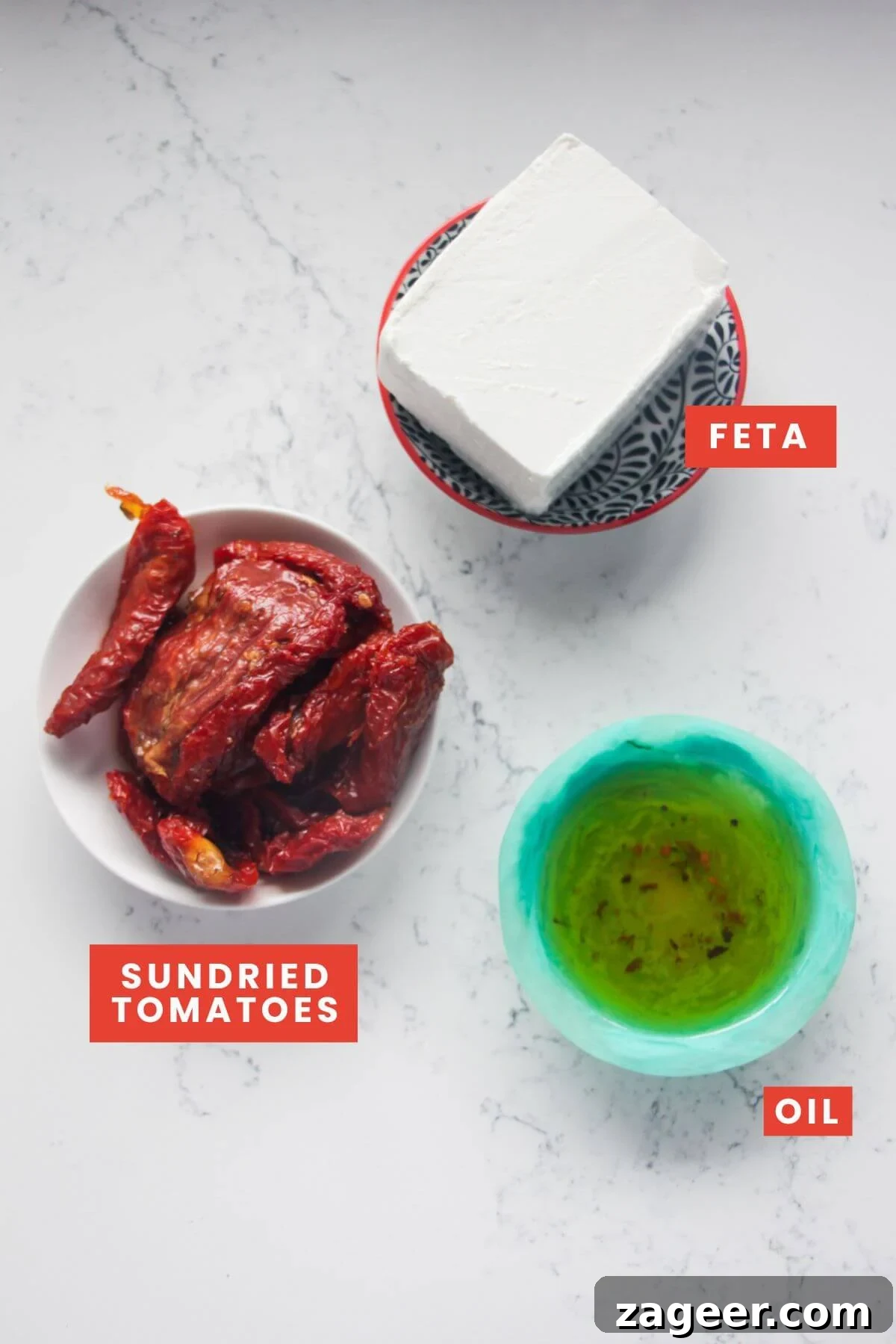 Instant Sun-Kissed Feta Dip 3 Ingredients for sundried tomato feta dip laid out on white marble background and labelled. A block of feta cheese, a jar of sundried tomatoes, and a small container of oil are visible, highlighting the simplicity of the recipe.