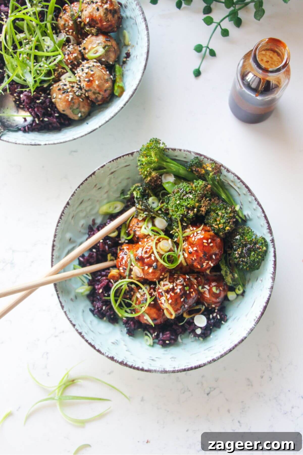 Sticky Gochujang Chicken Meatballs 2 Glazed Asian chicken meatballs with rice and broccoli in a small blue bowl, garnished with sesame seeds and scallions.