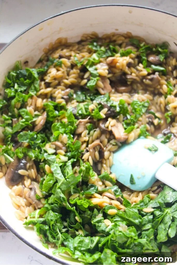 Velvety One-Pot Mushroom Orzo 9 Chopped spinach being stirred through mushroom orzo in a large white skillet.