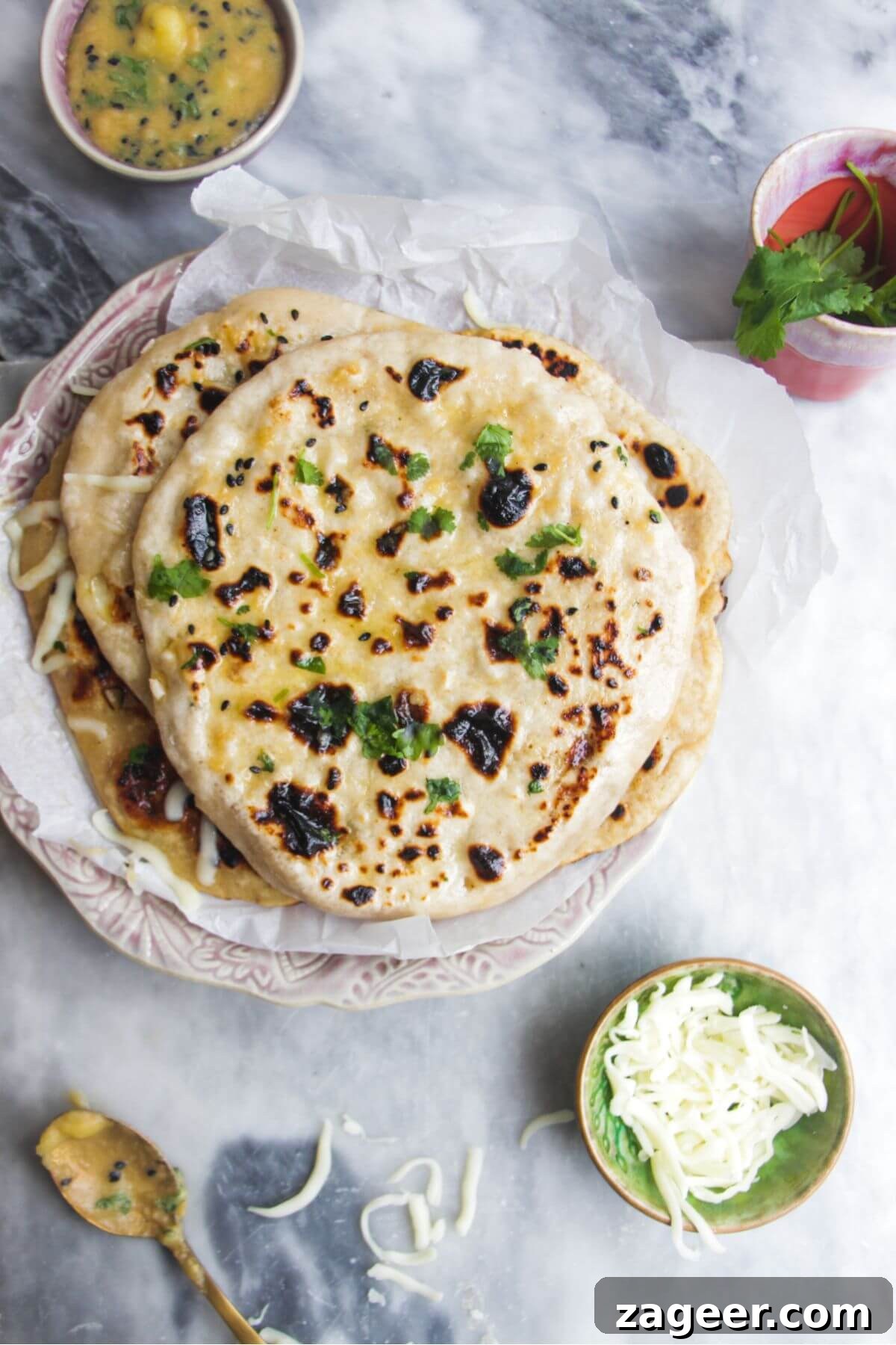 Effortless Two Ingredient Cheesy Naan 2 A stack of golden-brown cheese naan, oozing with melted cheese, piled on a small pink plate. A bowl of grated cheese and a pat of butter are nearby.