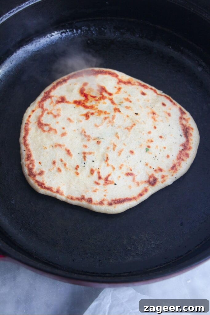 Effortless Two Ingredient Cheesy Naan 10 A golden, speckled cheese naan cooking in a large black cast iron skillet, with slight charring visible.