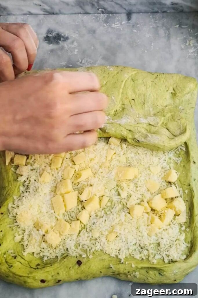 Wild Garlic & Cheddar Savory Hot Cross Buns 6 A pair of hands gently folds shredded cheese into the soft dough on a clean grey marble surface, demonstrating the incorporation process.