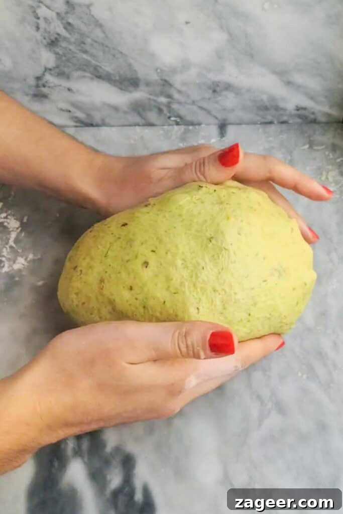 Wild Garlic & Cheddar Savory Hot Cross Buns 8 A pair of hands skillfully forms the large, cheese-infused dough into a smooth, round ball on a grey marble background.