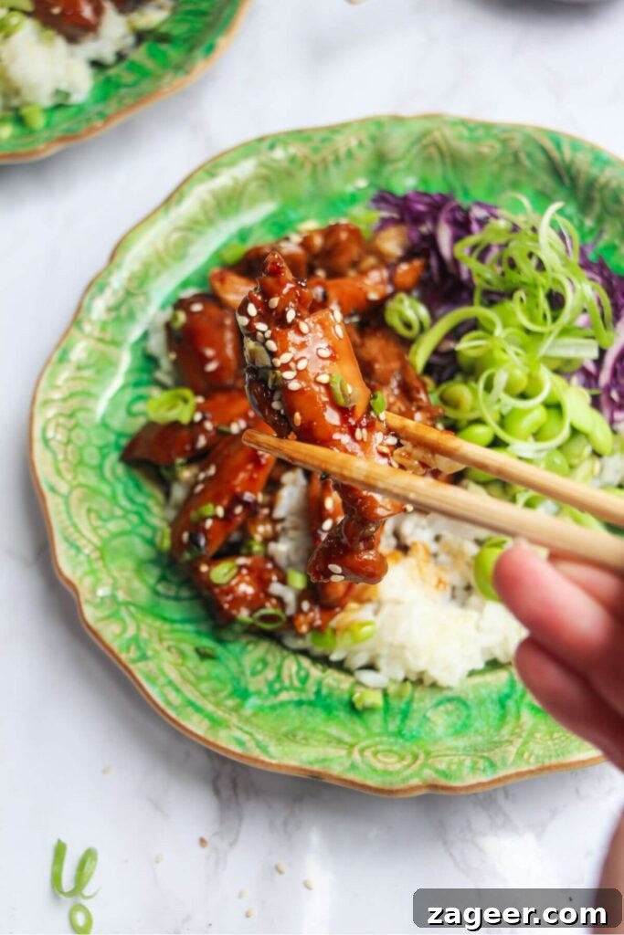 Hearty Teriyaki Chicken Rice Bowls 7 Hands holding chopsticks with a piece of teriyaki chicken, with a fully assembled teriyaki chicken bowl in the soft-focus background.