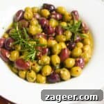 Close up of a white scalloped bowl of marinated olives, with skewers on the side.