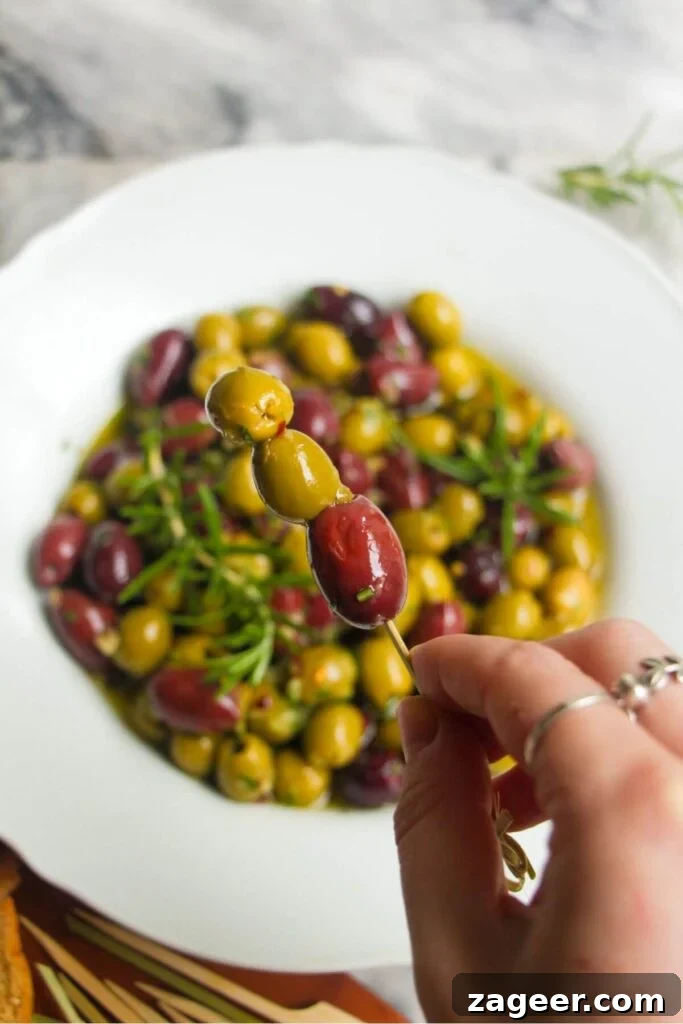 Zesty Olives in Minutes 7 A hand holding up three olives on a skewer with a bowl of marinated olives in the background, ready to enjoy.