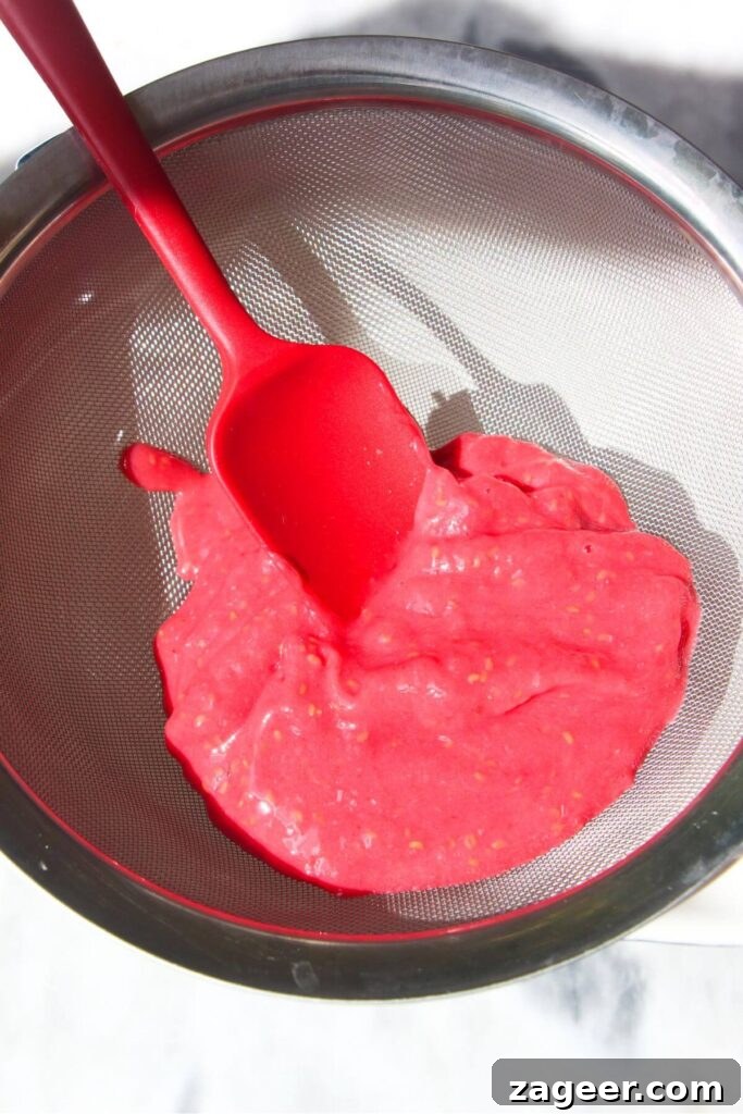 Heavenly Berry Ripple Cheesecake Pavlova Bites 4 Raspberry puree in a silver sieve with a red spatula inside.