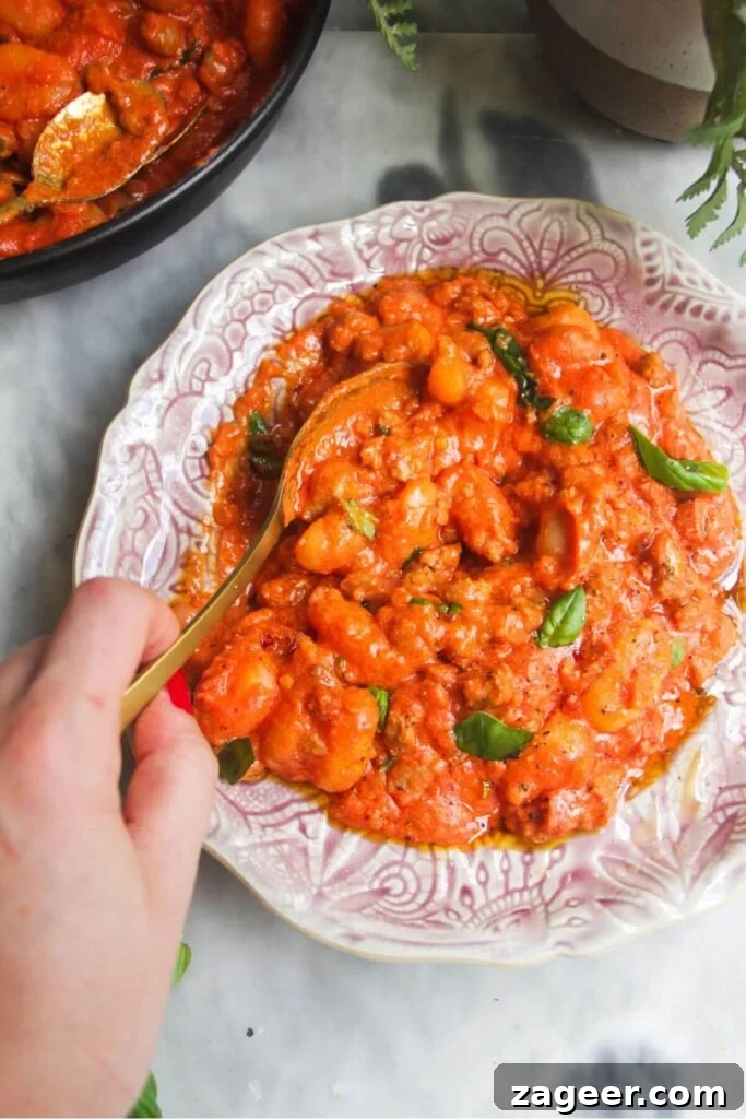 Creamy One-Pot Sausage Gnocchi Delight 11 An enticing close-up of creamy tomato sausage gnocchi on a pink plate, with a golden spoon ready to scoop. Fresh basil leaves add a pop of color and aroma.