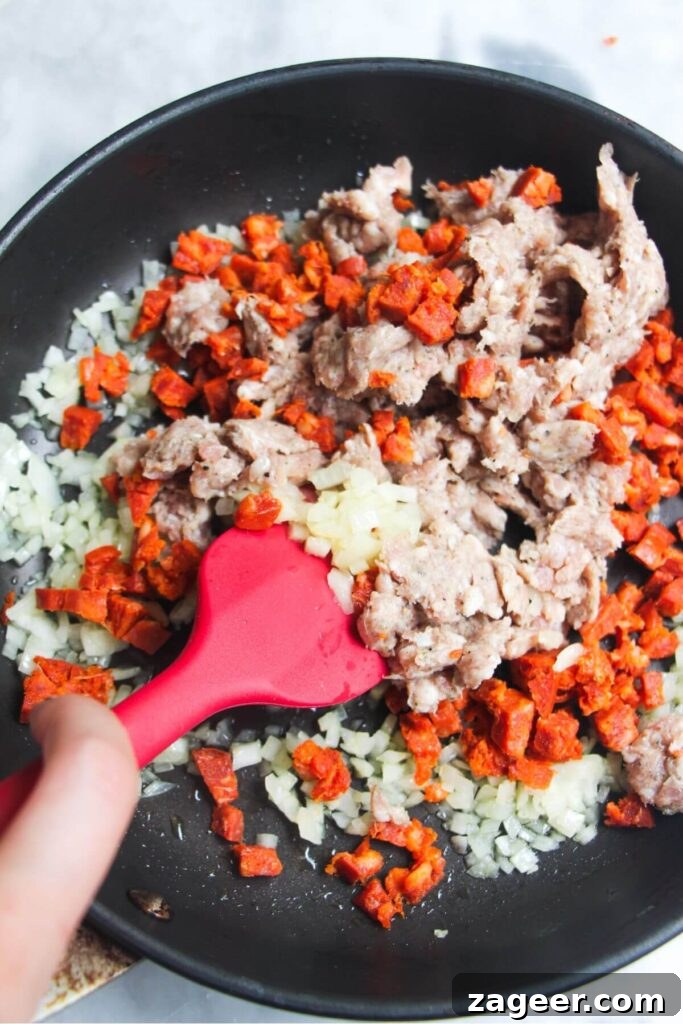 Creamy One-Pot Sausage Gnocchi Delight 4 A red spatula stirs finely diced onion and crumbled sausage meat in a high-sided pan, creating the savory base for the gnocchi dish.