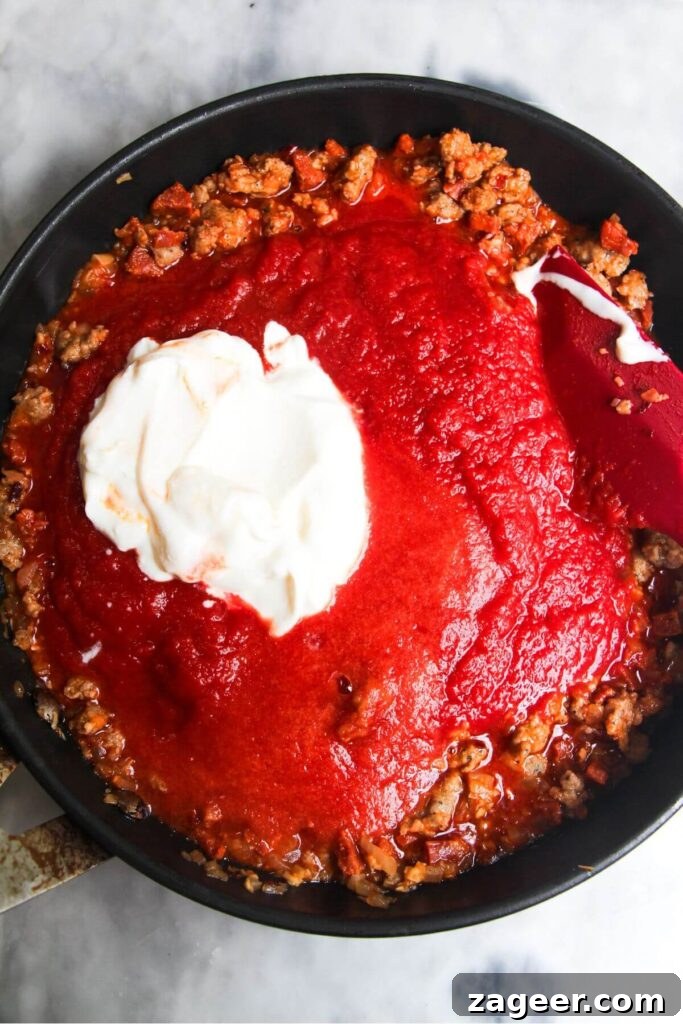 Creamy One-Pot Sausage Gnocchi Delight 6 Creamy sour cream and vibrant red tomato passata are being added to the cooked sausage mixture in the pan, forming the base of the luxurious sauce.