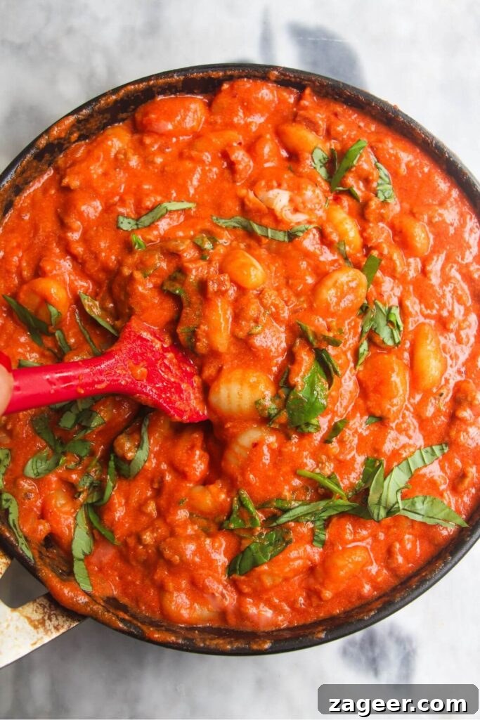 Creamy One-Pot Sausage Gnocchi Delight 9 A red spatula gently folds fresh basil leaves into the creamy tomato gnocchi, adding a burst of fresh flavor and color.