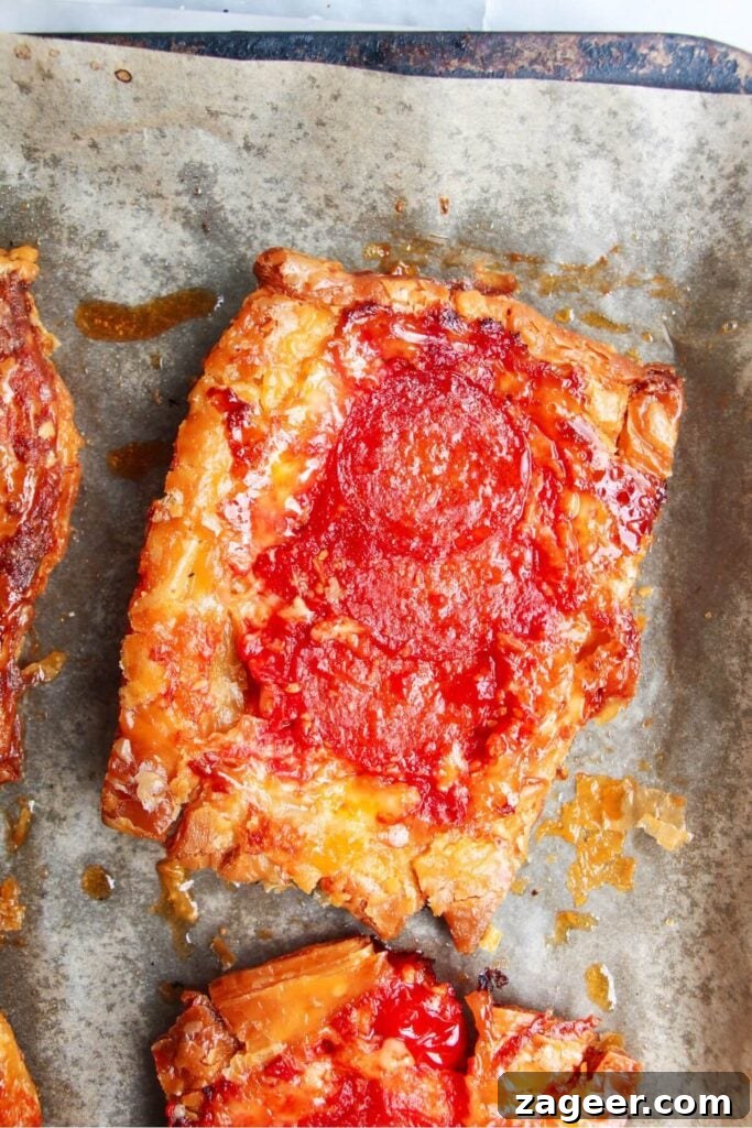 Puff Pastry Pizza Flips 9 A close-up shot of a single baked upside down puff pastry pizza tart, showcasing its bubbling cheese and crispy edges on a lined baking tray.