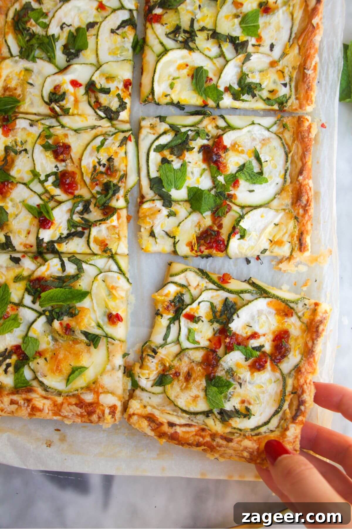 Golden Zucchini Puff Pastry Tart 2 A perfectly baked Zucchini Puff Pastry Tart, sliced into individual portions, with a hand gracefully removing one piece, showcasing its flaky crust and vibrant toppings.