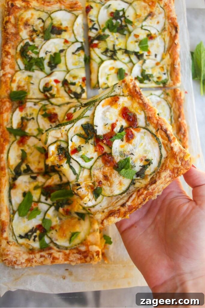 Golden Zucchini Puff Pastry Tart 9 A hand gently holds a single slice of the Zucchini Puff Pastry Tart, showcasing its delicious layers, with the remaining tart subtly blurred in the background.