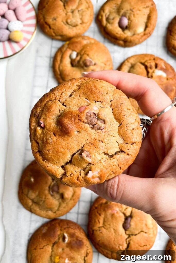A hand holding up a single, perfectly baked mini egg cookie, showcasing its soft center and crispy edges, with more cookies blurring in the background.