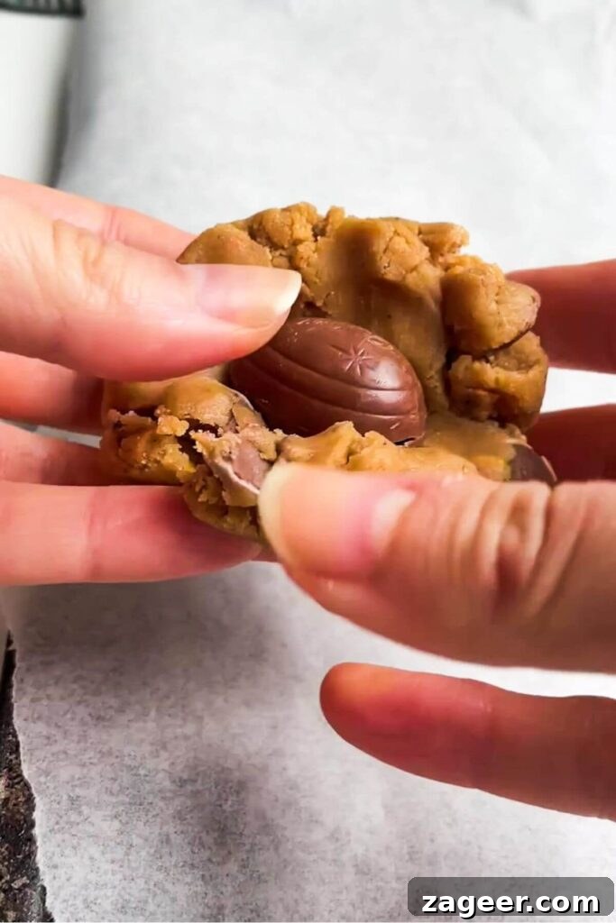 Hands meticulously folding the edges of the cookie dough around a mini Creme egg to encase it.