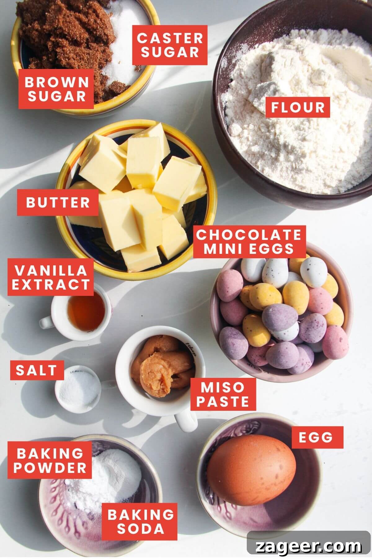 All the ingredients for mini egg cookies, including butter, sugars, miso paste, eggs, flour, and a bowl of colorful mini eggs, neatly laid out and labelled on a countertop.