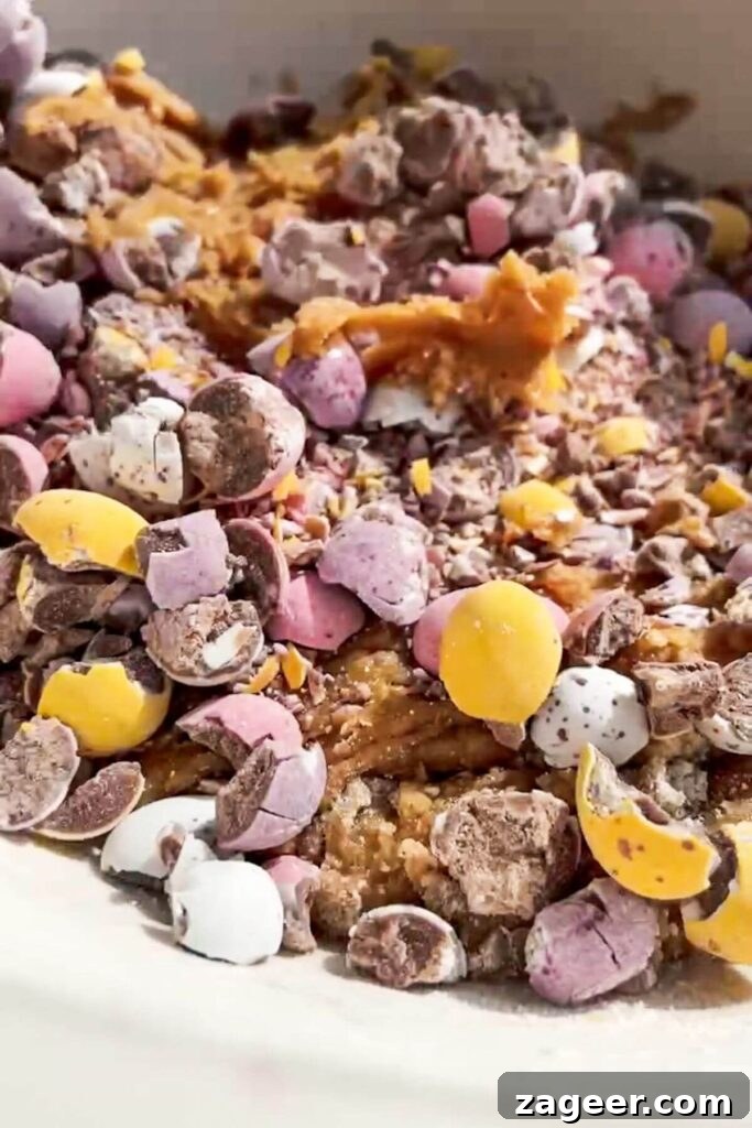 Crushed mini eggs being added to the cookie dough mixture in a large mixing bowl.
