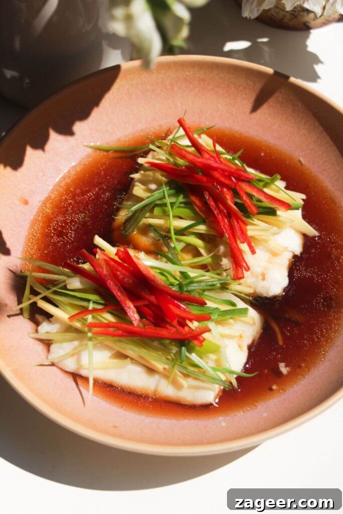 Aromatic Hot Oil Steamed Fish 9 Savory soy sauce dressing being poured generously over two steamed fish fillets, which are adorned with sliced scallions, chili, and ginger batons in a vibrant pink bowl.