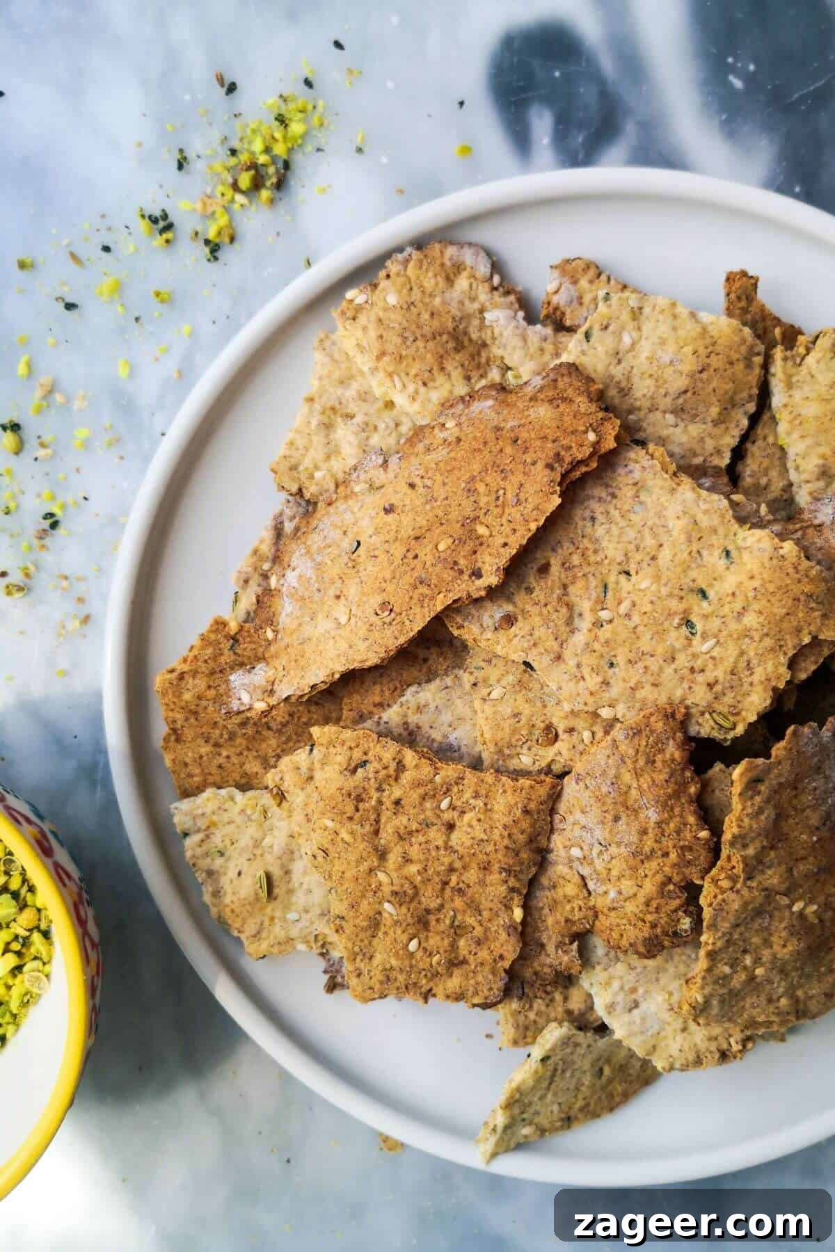 Tangy Sourdough Crisps with Aromatic Dukkah 2 Crispy sourdough discard crackers, generously seasoned with dukkah, arranged neatly on a small white plate. A small bowl of dukkah sits alongside, inviting a flavorful dip.