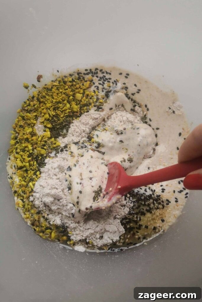 Tangy Sourdough Crisps with Aromatic Dukkah 3 All sourdough discard cracker ingredients mixed in a large clear bowl, being stirred with a red spatula.