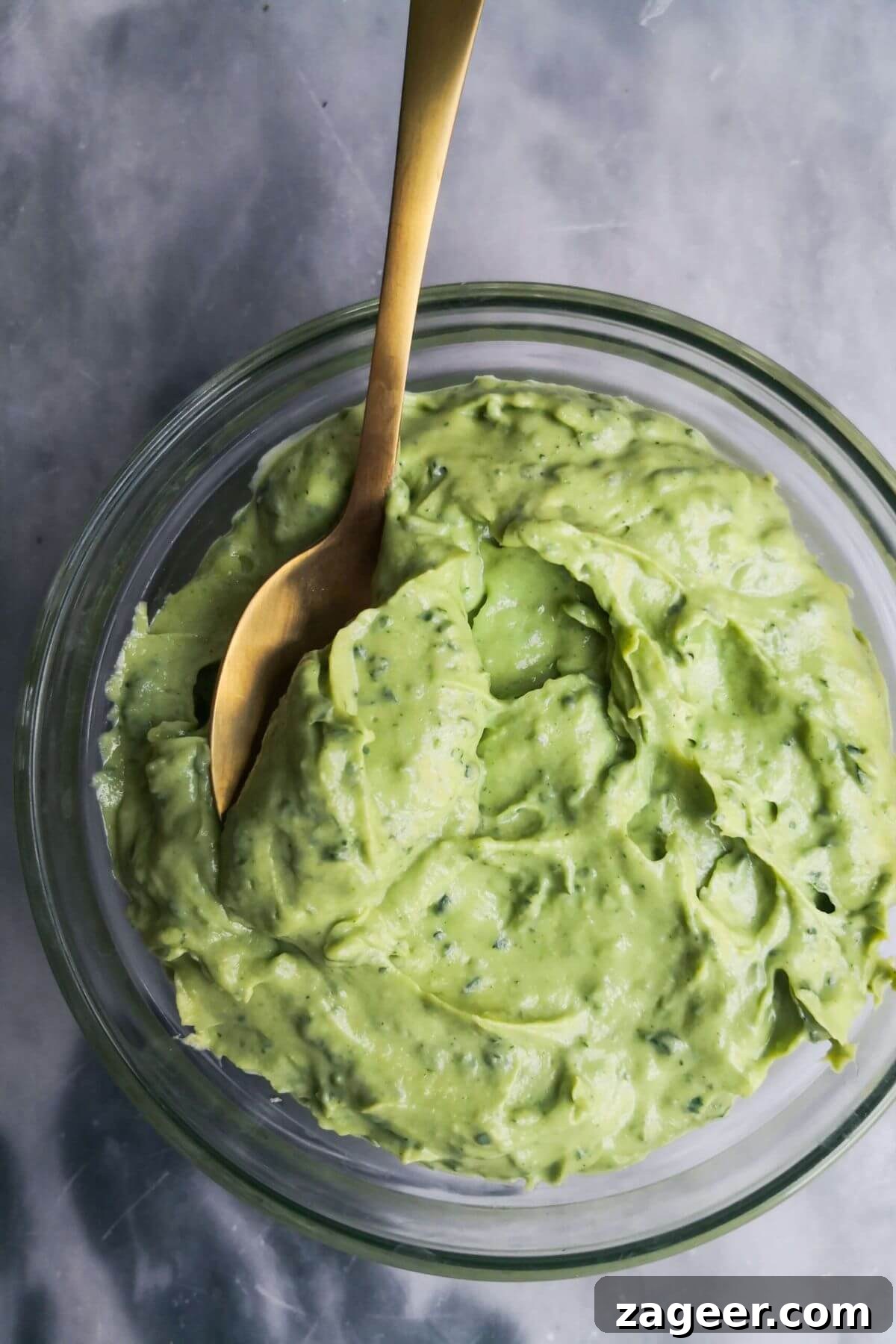 Vibrant 5-Minute Vegan Green Tahini Sauce 2 Green tahini in a small glass bowl with a gold spoon in it, on a grey marble background.
