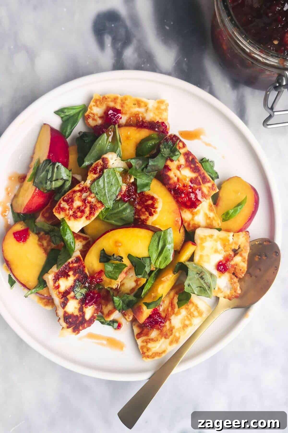 Sweet & Salty Peach Halloumi Salad with Chili Jam 2 A vibrant peach and halloumi salad artfully arranged on a pristine white serving plate, accompanied by a delicate small gold spoon. In the soft-focus background, a jar of sweet chili jam hints at the key ingredient, inviting a taste of summer.