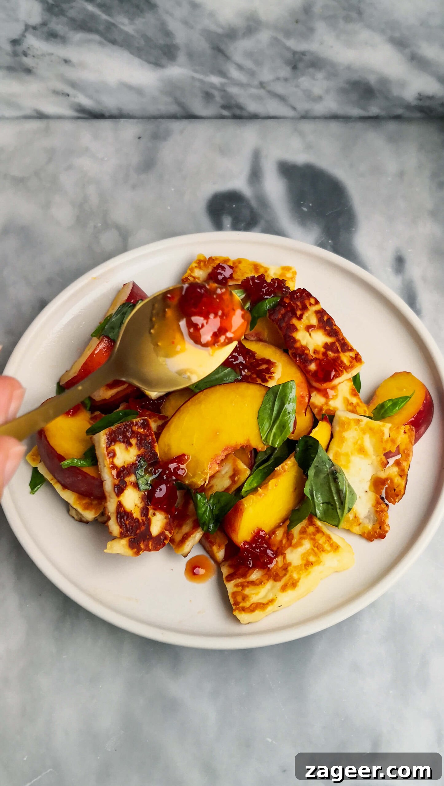 Sweet & Salty Peach Halloumi Salad with Chili Jam 9 A small gold spoon elegantly drizzling sweet chili jam onto a freshly prepared peach and halloumi salad, set against a chic grey marble background, ready for serving.