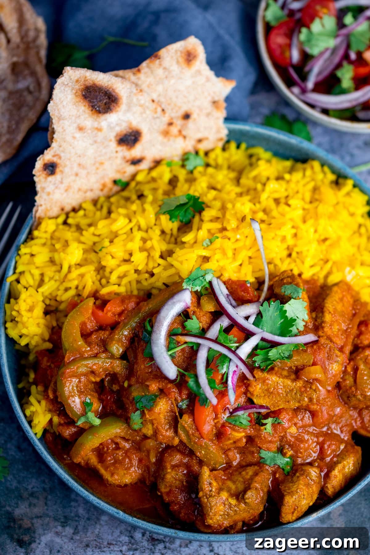 Vibrant Spicy Chicken Jalfrezi 3 Overhead shot of a blue bowl filled with homemade Chicken Jalfrezi and fluffy pilau rice, garnished with finely chopped red onion and fresh coriander. A piece of chapati is visible on the edge of the bowl.
