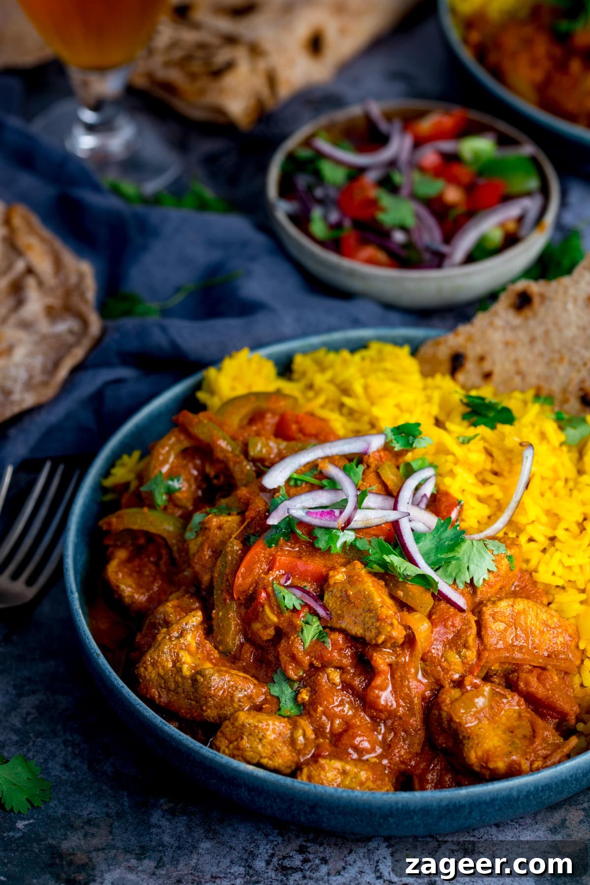 Vibrant Spicy Chicken Jalfrezi 8 A vibrant spread of Chicken Jalfrezi and pilau rice in a blue bowl, accompanied by a refreshing tomato and onion salad, chapatis, and forks on a complementary blue background.
