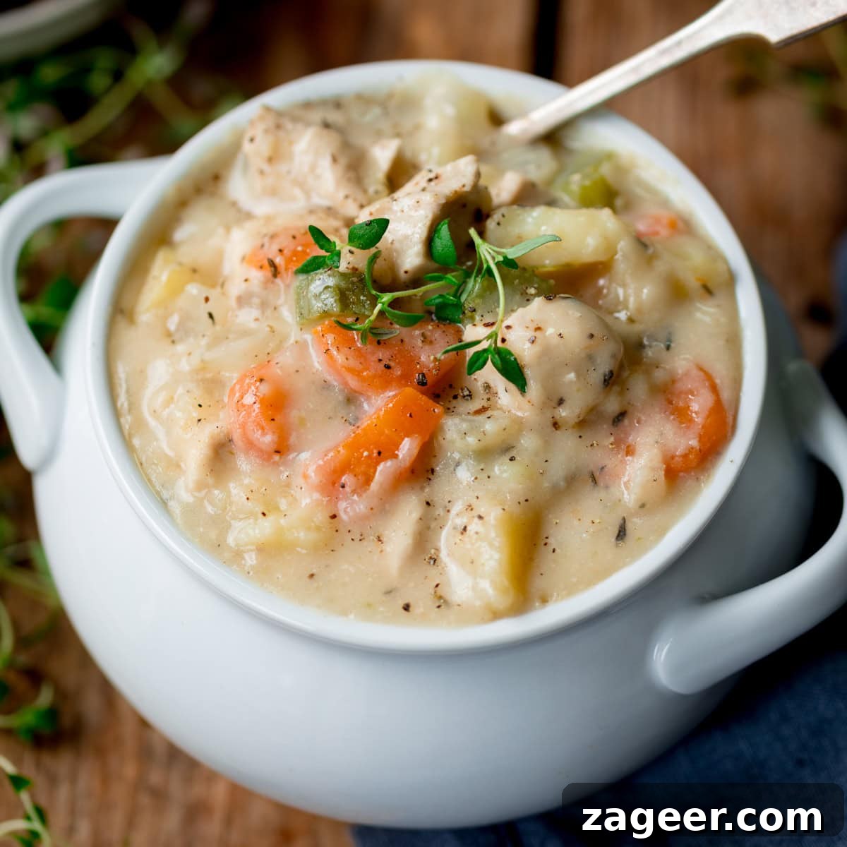 Creamy Chicken Pot Pie Comfort 2 A white bowl filled with creamy chicken pot pie soup containing pieces of chicken, carrots, celery and potatoes, topped with fresh thyme, invitingly placed on a rustic wooden surface with a spoon.