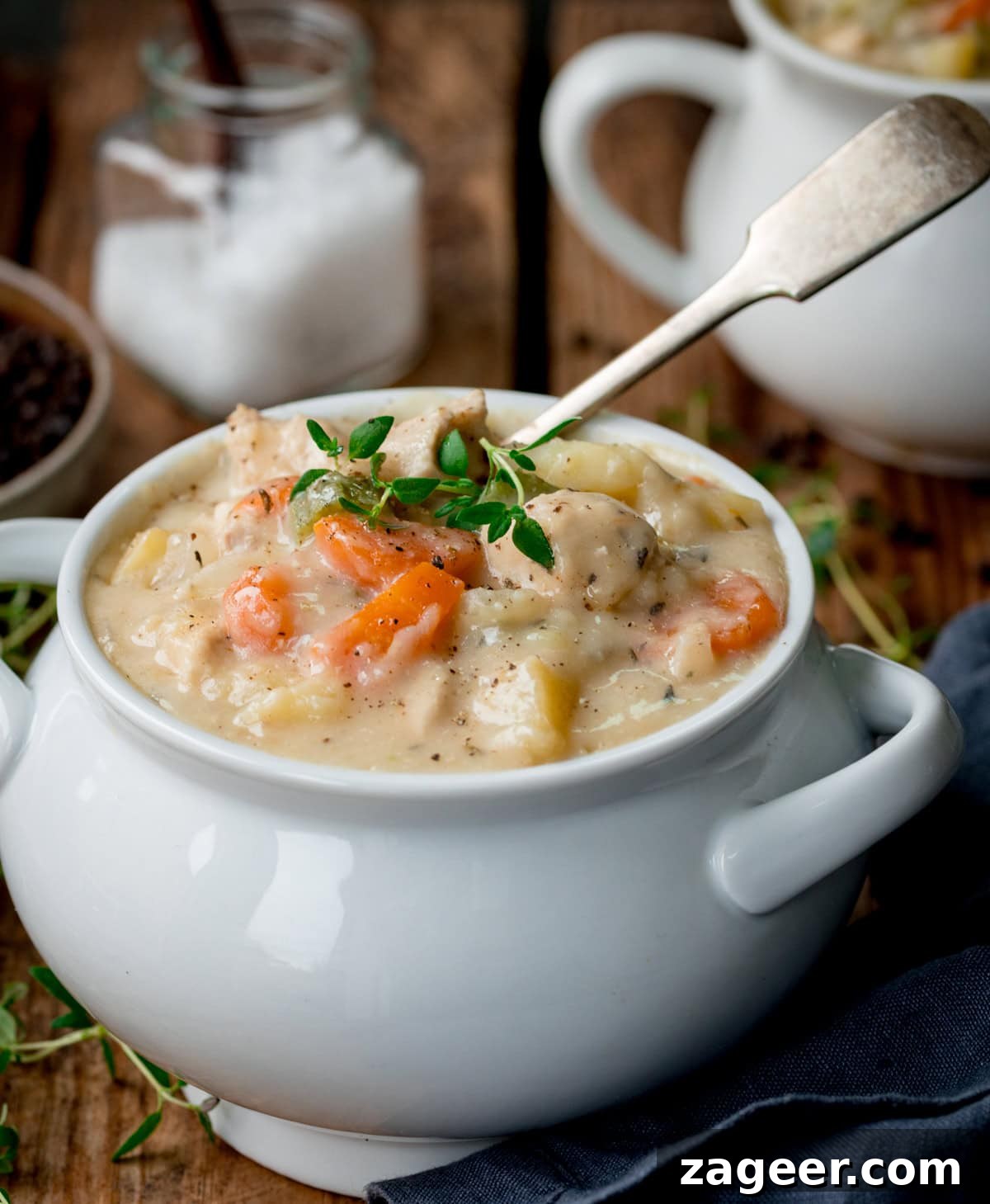 Creamy Chicken Pot Pie Comfort 4 A large, rustic white bowl filled with creamy chicken pot pie soup, garnished with fresh thyme leaves. A vintage silver spoon rests in the bowl, while other bowls and kitchen accessories are blurred in the background on a wooden table. The lighting is warm and inviting.