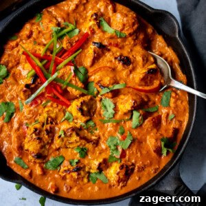 A frying pan filled with Chicken Tikka Masala, garnished with coriander and sliced red chilli, with a spoon resting inside.