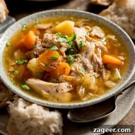Square image of a steaming bowl of chicken and vegetable soup in a light bowl, elegantly placed on a plate. A rustic chunk of bread and a spoon are nestled invitingly between the bowl and plate, ready for a comforting meal.