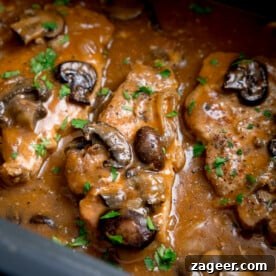 Square close-up image of smothered pork chops with tender mushrooms and a thick, savory gravy in the pan of a slow cooker. Chopped parsley is delicately sprinkled on top for freshness.