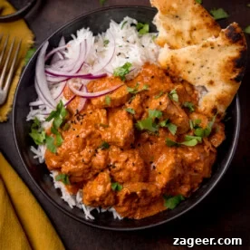 Overhead picture of a dark bowl full of fluffy boiled rice and flavorful crock pot butter chicken, garnished with some sliced red onion and charred naan bread tucked into the corner of the bowl.