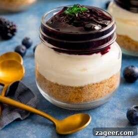 An individual berry cheesecake, perfectly layered in a mason jar, presented on a vibrant blue background next to a gleaming gold teaspoon, highlighting its delicious and portable nature.
