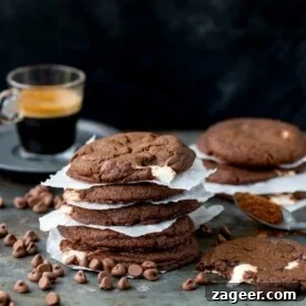 Close-up of a stack of rich double chocolate nougat cookies, with baking paper between each cookie. In the foreground are chocolate chips and a cookie with a bite taken out, while a glass of espresso and another stack of cookies are visible in the background.