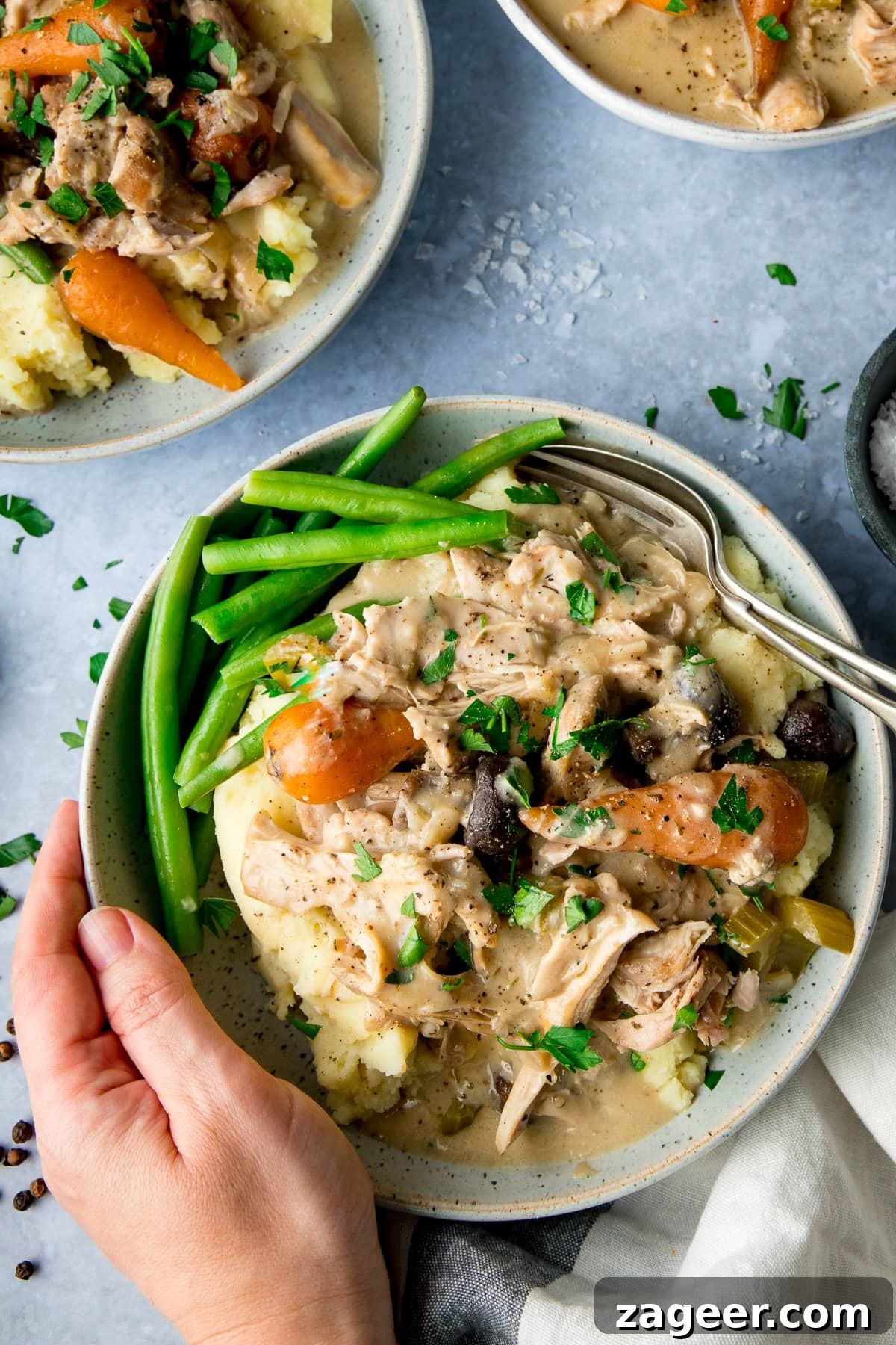 Creamy Chicken Casserole Meal A hand holding a bowl of creamy chicken casserole with mashed potatoes and green beans, against a light blue background.